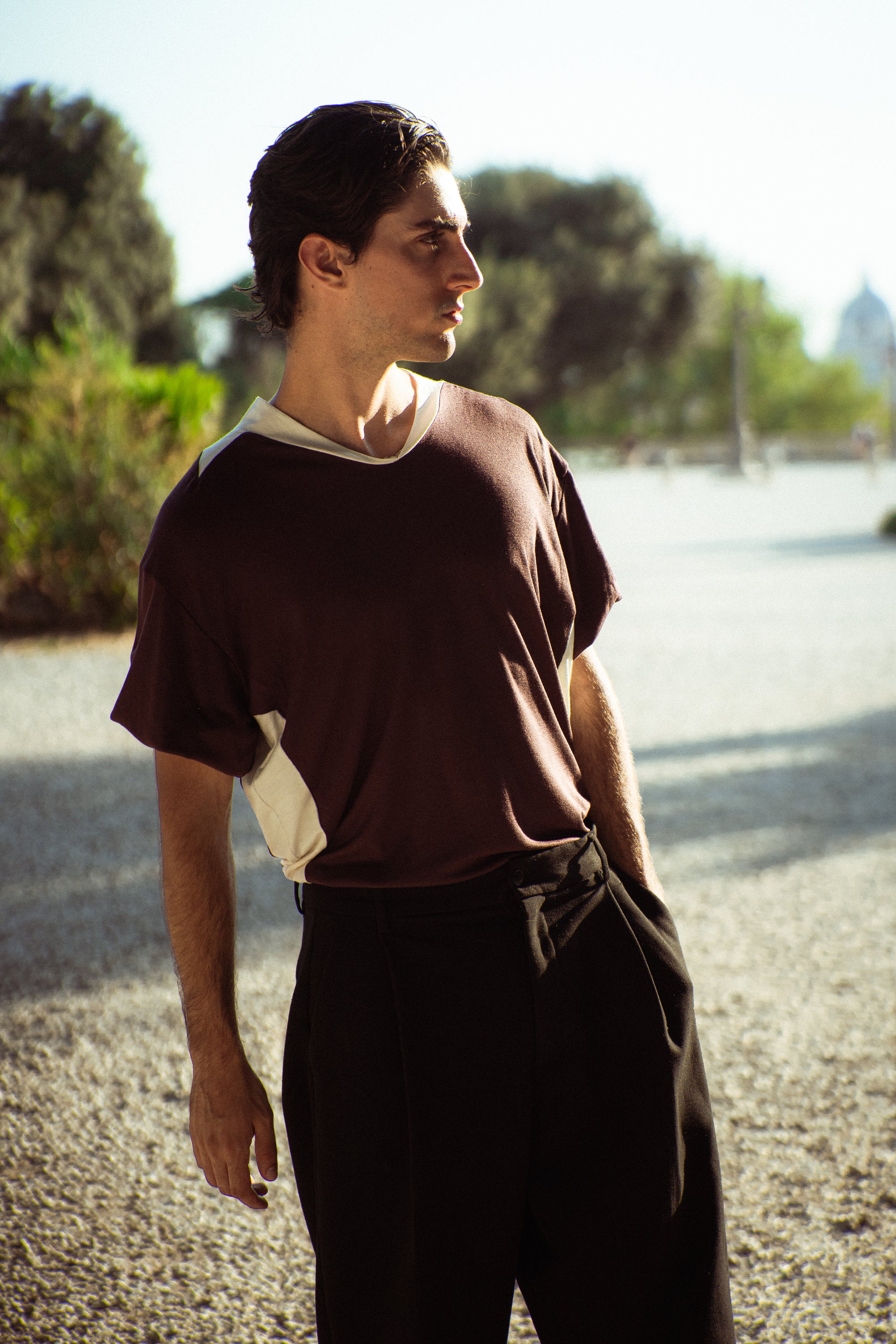 A man wearing a dark brown and cream color-blocked t-shirt and black pants, standing outdoors on a gravel path with trees in the background.