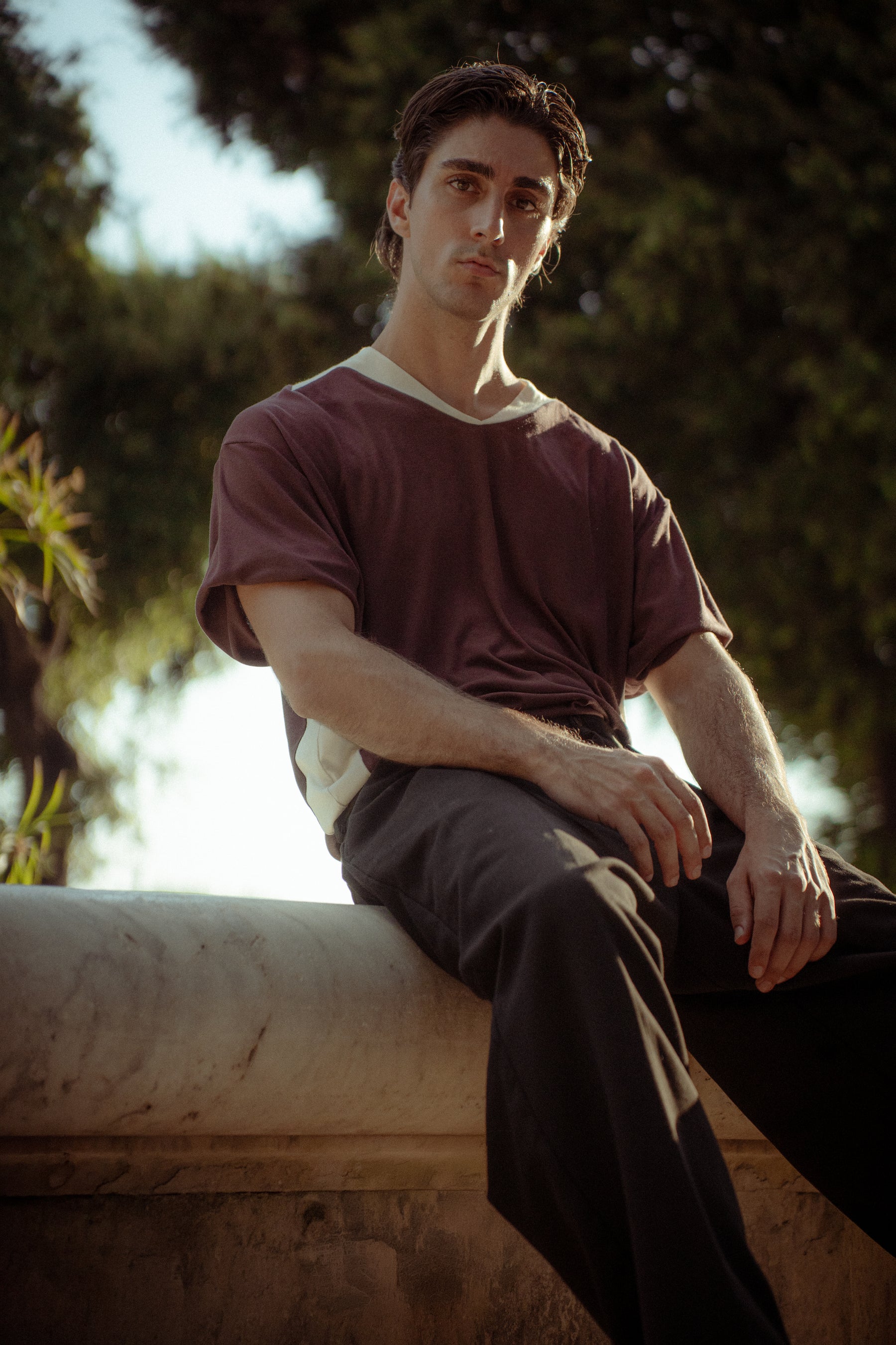 A man sitting outdoors on a stone ledge, wearing a brown short-sleeve t-shirt with a white crew neck and dark trousers.