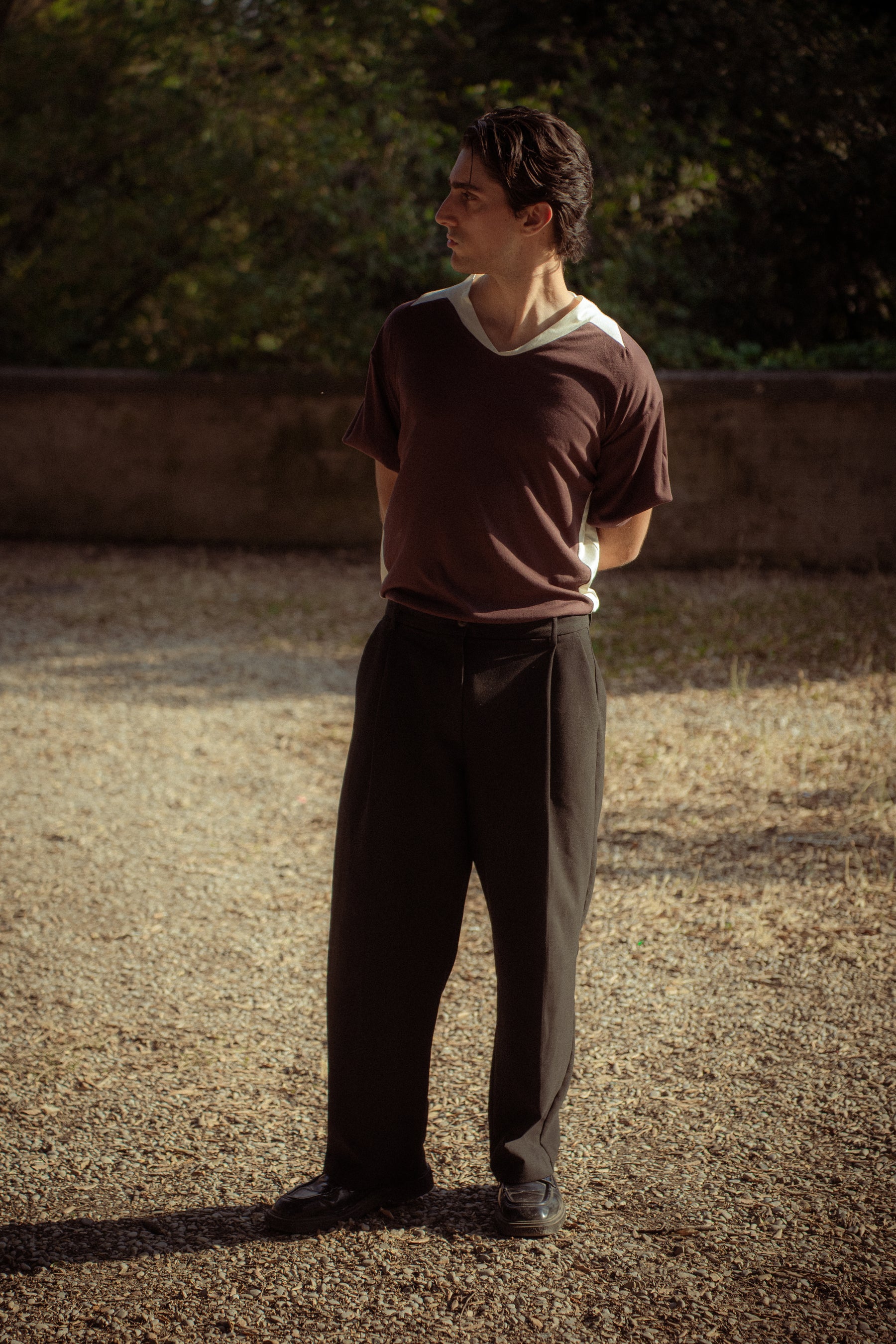 A man standing outdoors on a gravel path wearing a dark brown short-sleeve t-shirt with a white v-neck collar,  black trousers, and black shoes.