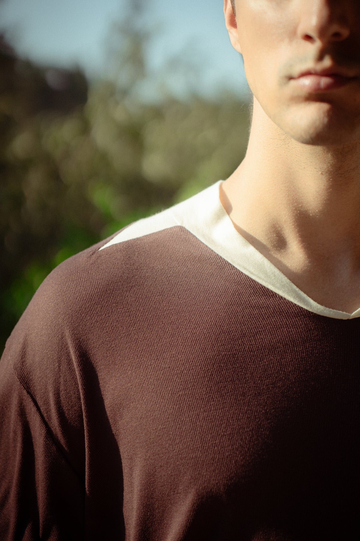 A close-up lifestyle shot of a man wearing a brown long-sleeve knit shirt with a contrasting cream ribbed v-neck collar, set against a blurred outdoor background.