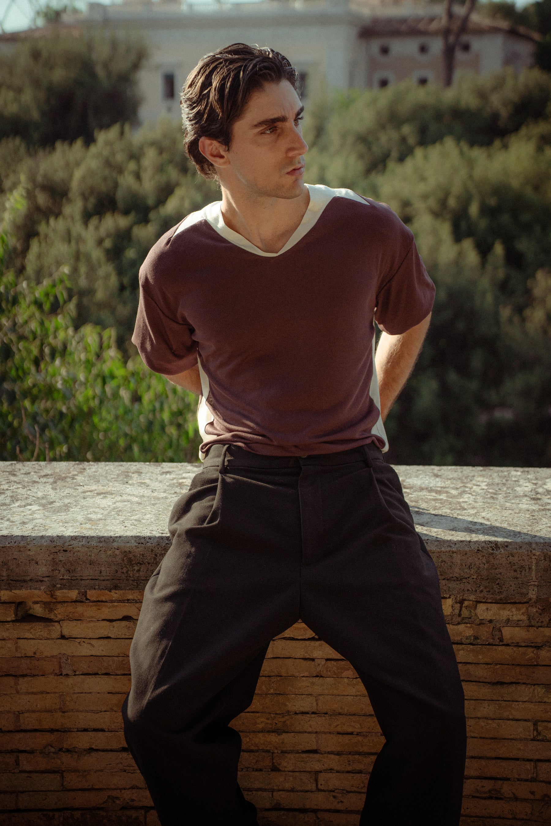 A man wearing a brown v-neck t-shirt with a cream collar and dark trousers, sitting on a stone wall with a green outdoor background.